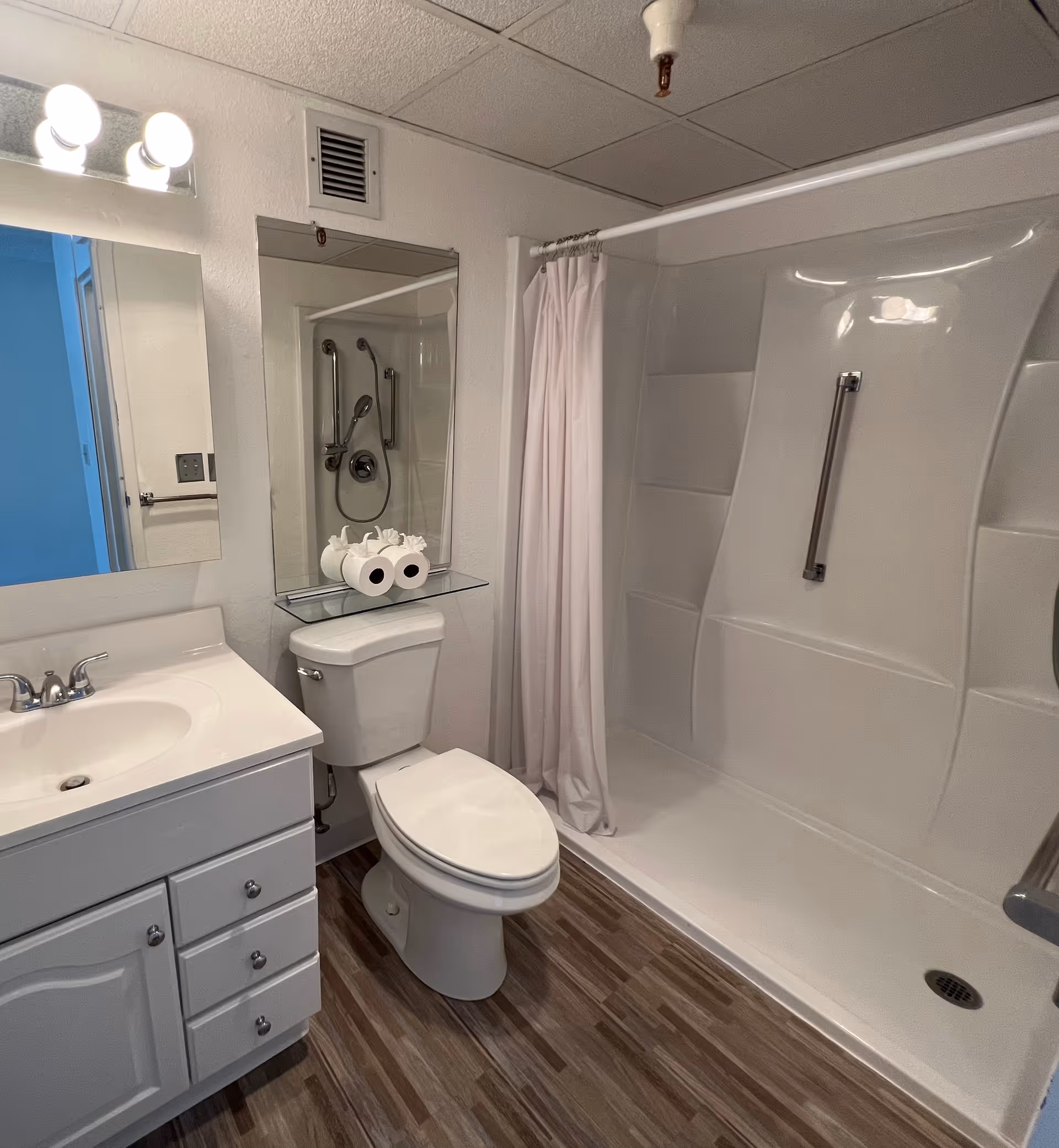 A clean bathroom with a white sink and vanity on the left, a toilet in the center with two rolls of toilet paper on a glass shelf above it, and a white shower with a curtain and grab bar on the right. The floor has wood-like tiles and the walls are white.