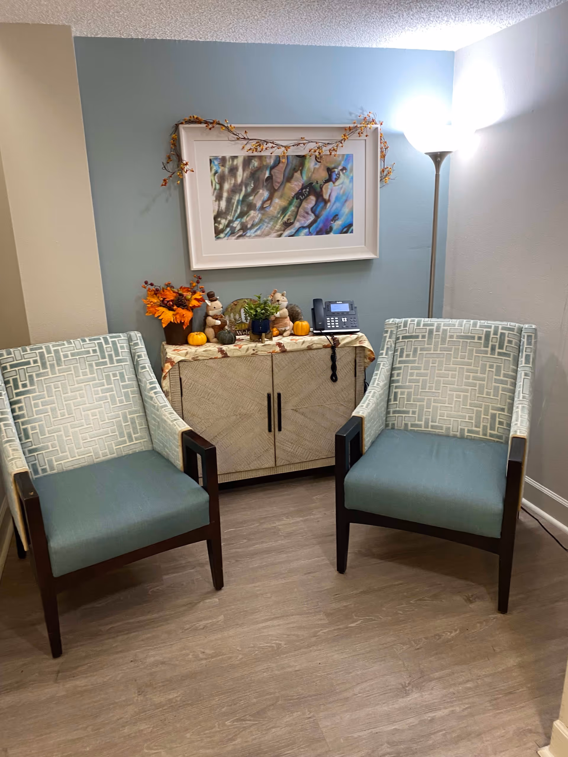 A cozy corner with two patterned armchairs facing forward, a wooden cabinet between them decorated with small pumpkins, autumn-themed figurines, a plant, and a telephone. Above the cabinet is a framed abstract artwork with a garland draped over it. A tall floor lamp is positioned to the right, illuminating the space.