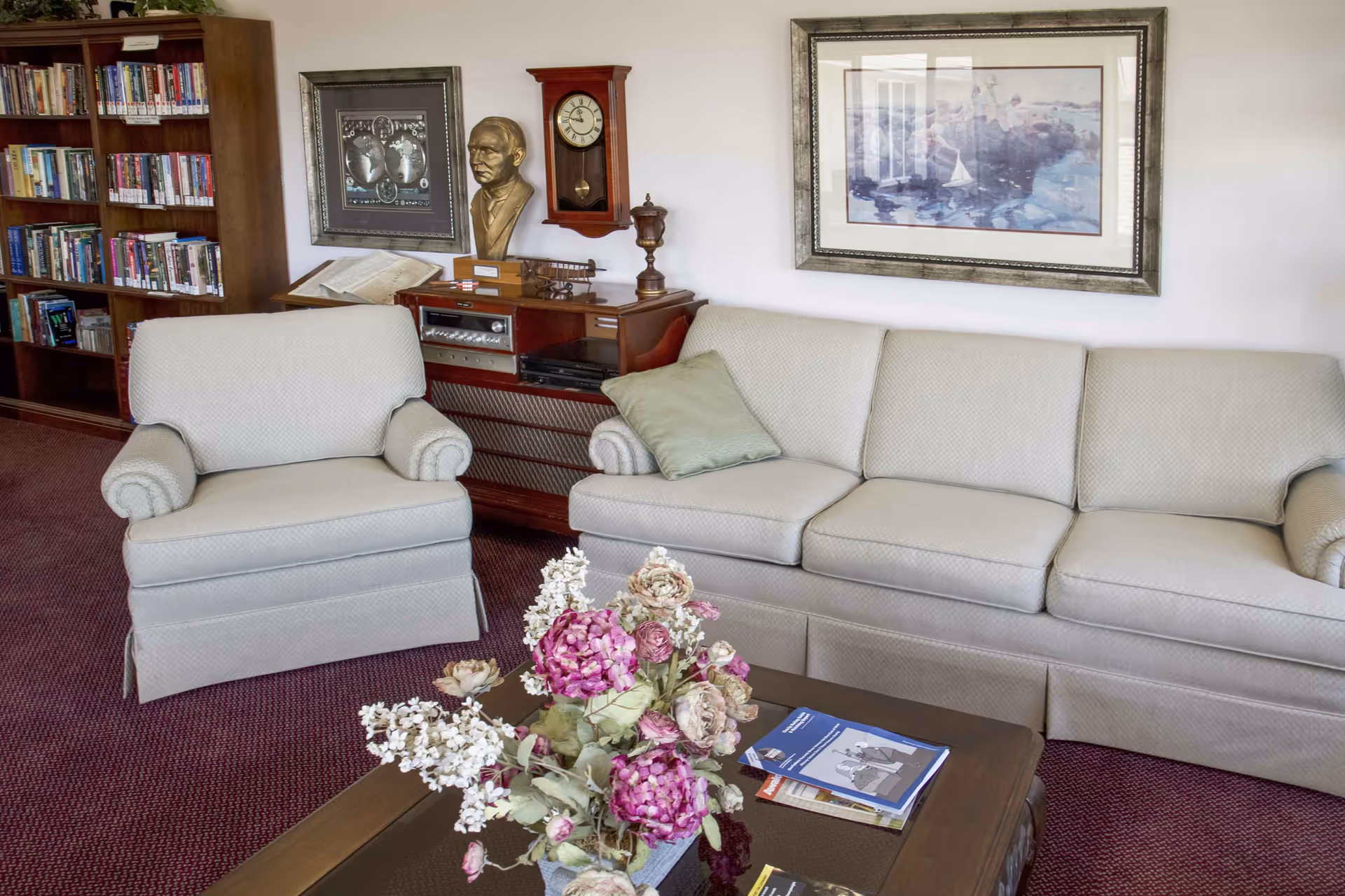 A cozy living room area with a beige armchair and matching sofa, a wooden coffee table with a floral arrangement and magazines, a wooden bookshelf filled with books, a vintage-style clock, a bust sculpture, and framed artwork on the walls.