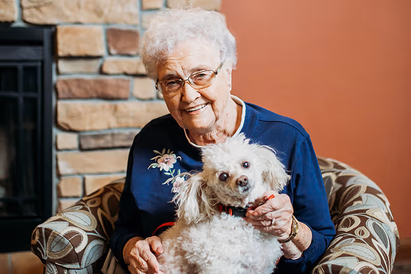 An elderly woman sitting in an upholstered chair holding a small white poodle in a cozy living room by a stone fireplace.