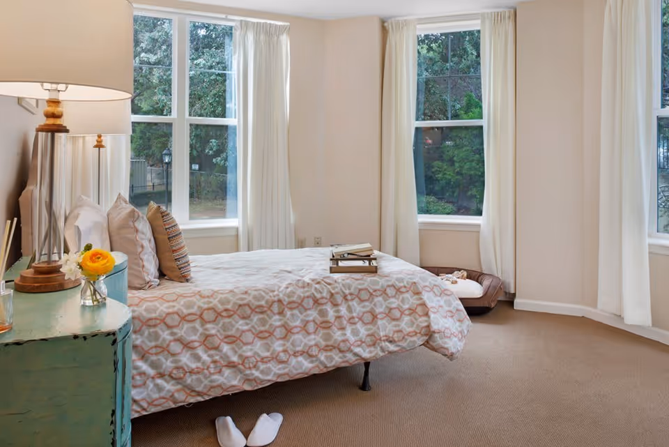 A bright bedroom with a bed covered in a patterned duvet and several pillows. There is a small turquoise nightstand with a lamp, a small vase with flowers, and other small items. Three large windows with white curtains let in natural light and show greenery outside. A pair of white slippers is on the carpeted floor near the bed, and a pet bed is placed near one of the windows.