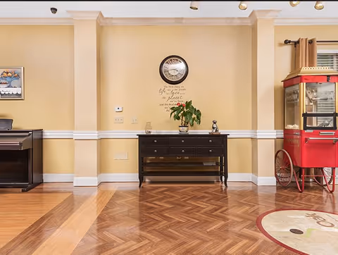 Bright common area with a wooden console table under a wall clock, a plant on the table, a piano to the left and a red popcorn machine to the right on a parquet floor.