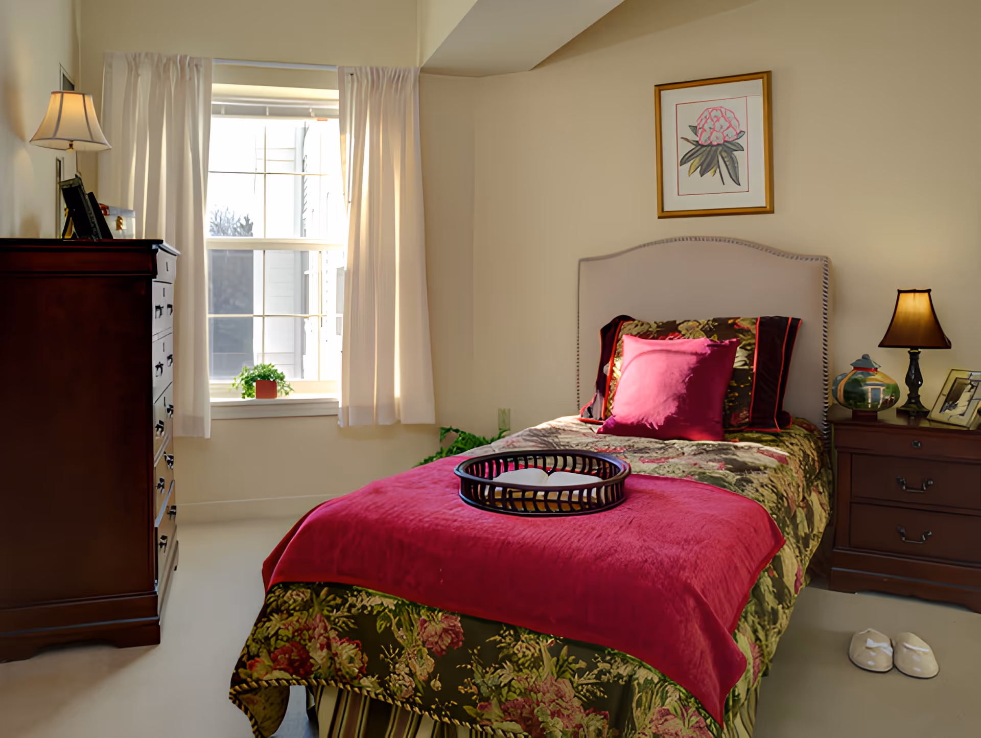 Sunlit single bedroom with floral bedding and a magenta throw, a nightstand with lamp, dresser, and a window with curtains.