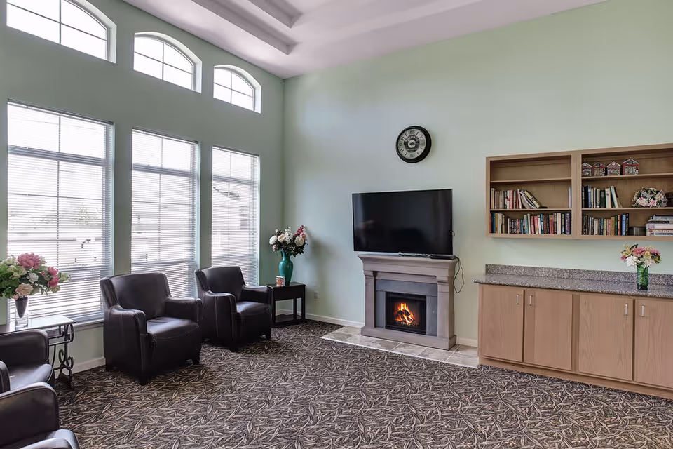 A cozy living room area with three black leather armchairs arranged near large windows with blinds. There is a fireplace with a TV mounted above it, a wall clock above the TV, and a built-in wooden bookshelf and cabinet on the right side. The room has light green walls and patterned carpet flooring, with flower vases placed on side tables and the cabinet.