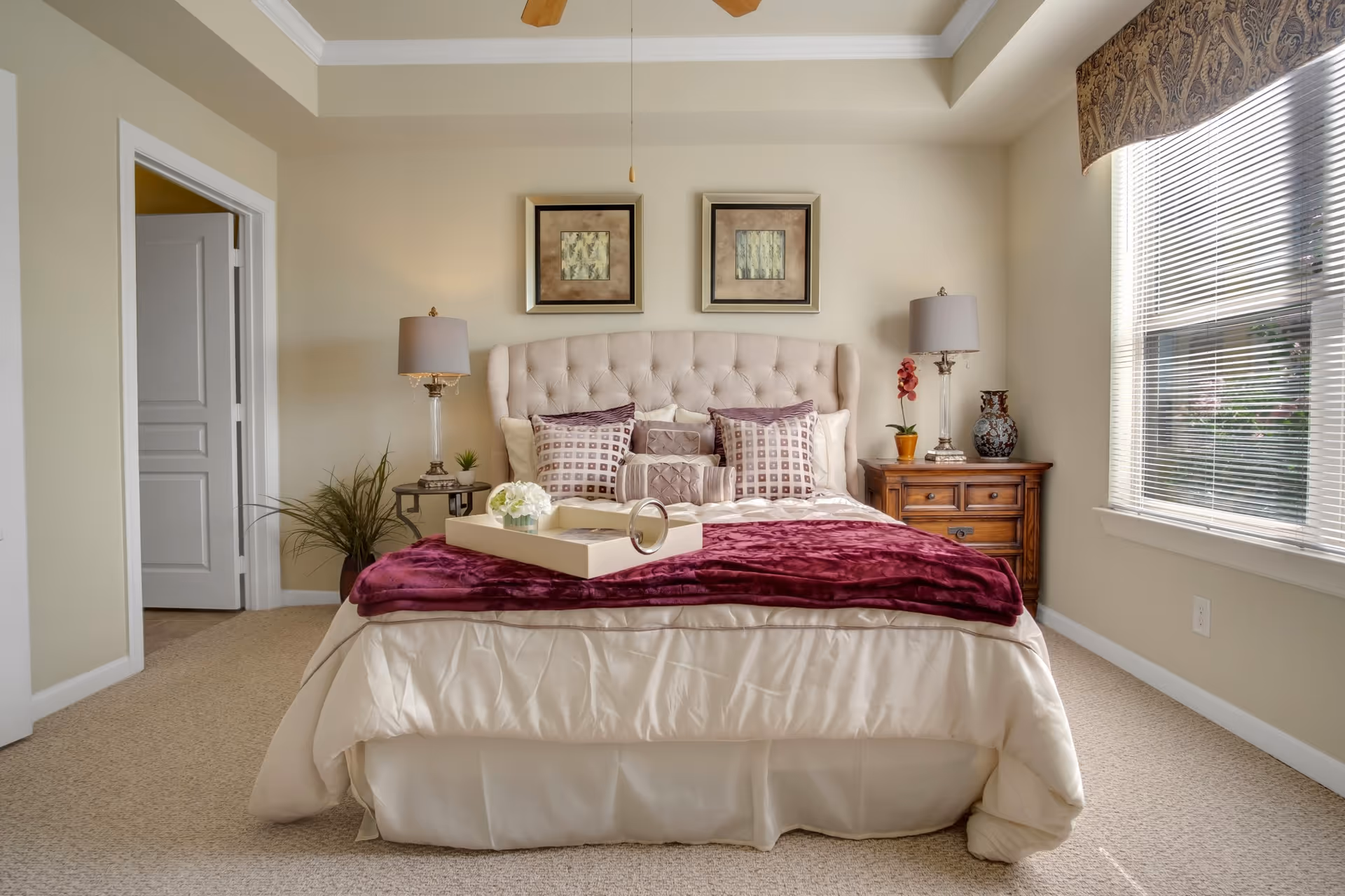 A neatly made bed with a tufted headboard in a bedroom. The bed has cream-colored bedding with a burgundy throw blanket and multiple decorative pillows. There are two framed artworks above the headboard. On each side of the bed are wooden nightstands with table lamps, a small plant, and decorative items. A large window with blinds and a valance allows natural light into the room. The walls are painted beige and the carpet is light-colored. An open doorway leads to another room.
