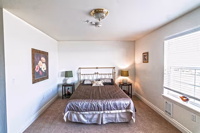 A simple bedroom with a metal bed frame and a brown bedspread. There are two bedside tables, each with a green lamp. A window with blinds is on the right wall, and two framed pictures hang on the walls. The room has beige carpet and white walls.