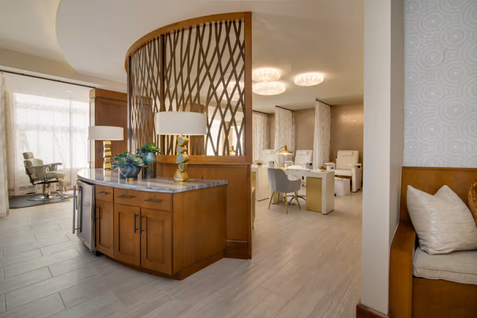 Interior view of a modern, well-lit room with a wooden cabinet topped with a marble surface and decorative lamps and plants. Behind a decorative wooden partition, there are several white chairs and tables arranged in a salon or spa-like setting. To the right, there is a cushioned bench with pillows against a patterned wall.
