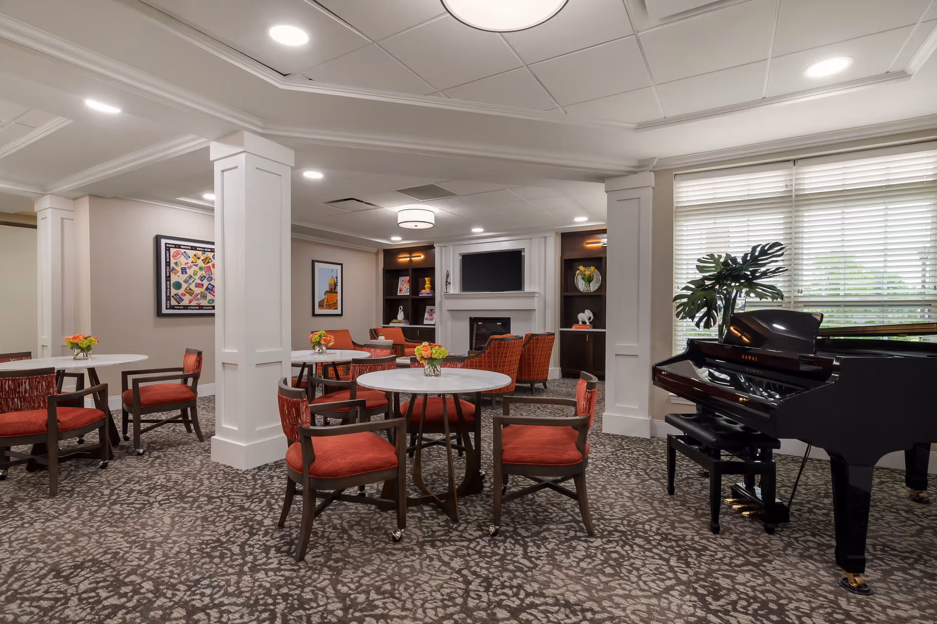 A cozy common area with round tables and red cushioned chairs, a black grand piano near a large window with blinds, a fireplace with a TV above it, and built-in shelves with decorative items. The room has patterned carpet and soft lighting.