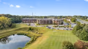 A wide aerial view of The Summit senior living facility showing a large multi-story building surrounded by green lawns, a small pond in the foreground, and a parking lot with several cars. Trees and a clear blue sky are visible in the background.