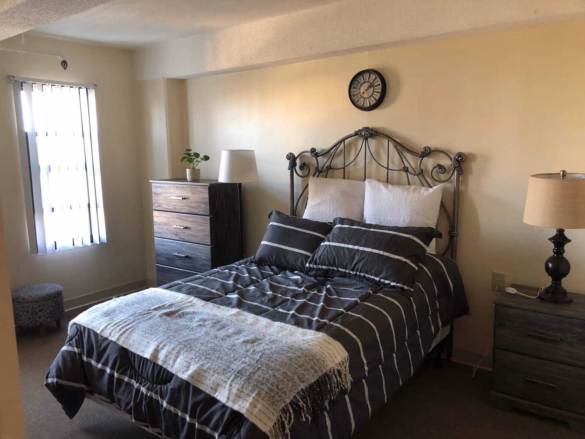 A furnished bedroom with a double bed with striped bedding, two bedside tables and lamps, a dresser, a window with vertical blinds, and a wall clock.