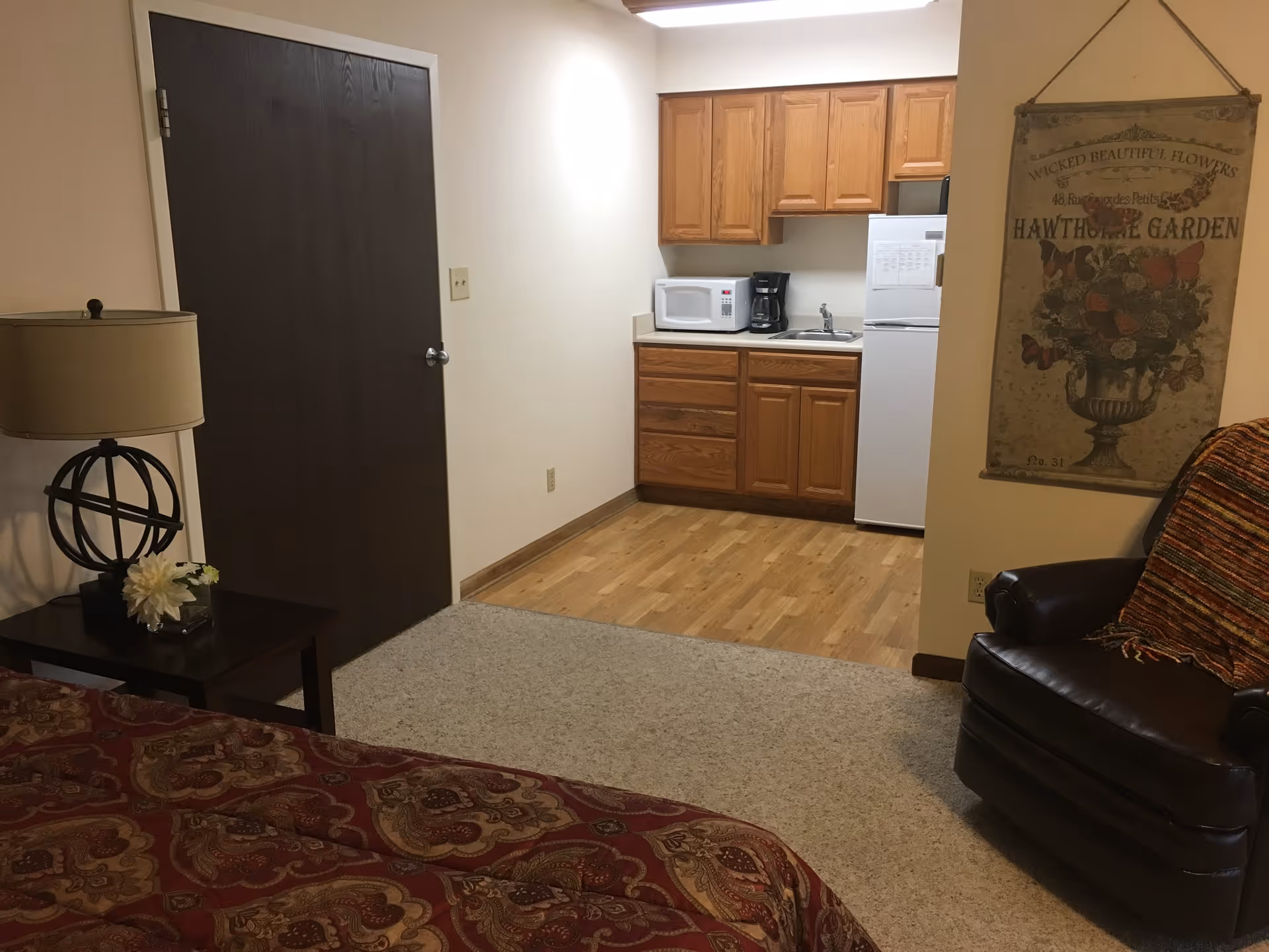 A small living space featuring a kitchenette with wooden cabinets, a white microwave, coffee maker, sink, and refrigerator. In the foreground, there is a bed with a patterned red bedspread, a side table with a lamp and flower decoration, and a dark brown leather armchair with a colorful throw blanket. A decorative wall hanging with floral and butterfly designs is visible on the right wall.