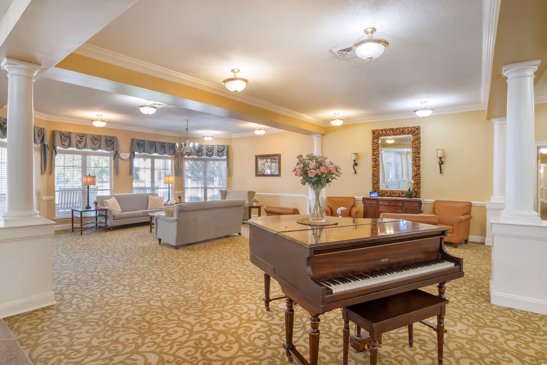 A spacious and well-lit living room area with beige patterned carpet, cream-colored walls, and white columns. The room features a dark wooden grand piano with a vase of pink flowers on top, several seating areas with gray sofas and tan armchairs, side tables with lamps, a large ornate mirror on the wall, and large windows with gray valances allowing natural light to enter.