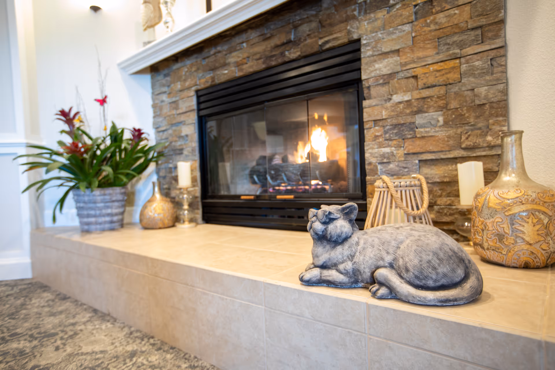 A cozy indoor fireplace with a stone surround and a lit fire. On the tiled hearth in front of the fireplace, there is a decorative gray cat statue wearing glasses, a large patterned vase, a candle, a wicker lantern, and a potted plant with green leaves and red flowers. The setting appears warm and inviting.