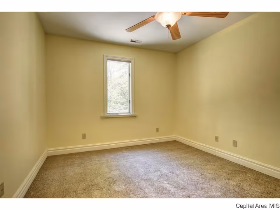 Empty room with beige walls, a single window, beige carpet flooring, white baseboards, and a ceiling fan with wooden blades and a light fixture.
