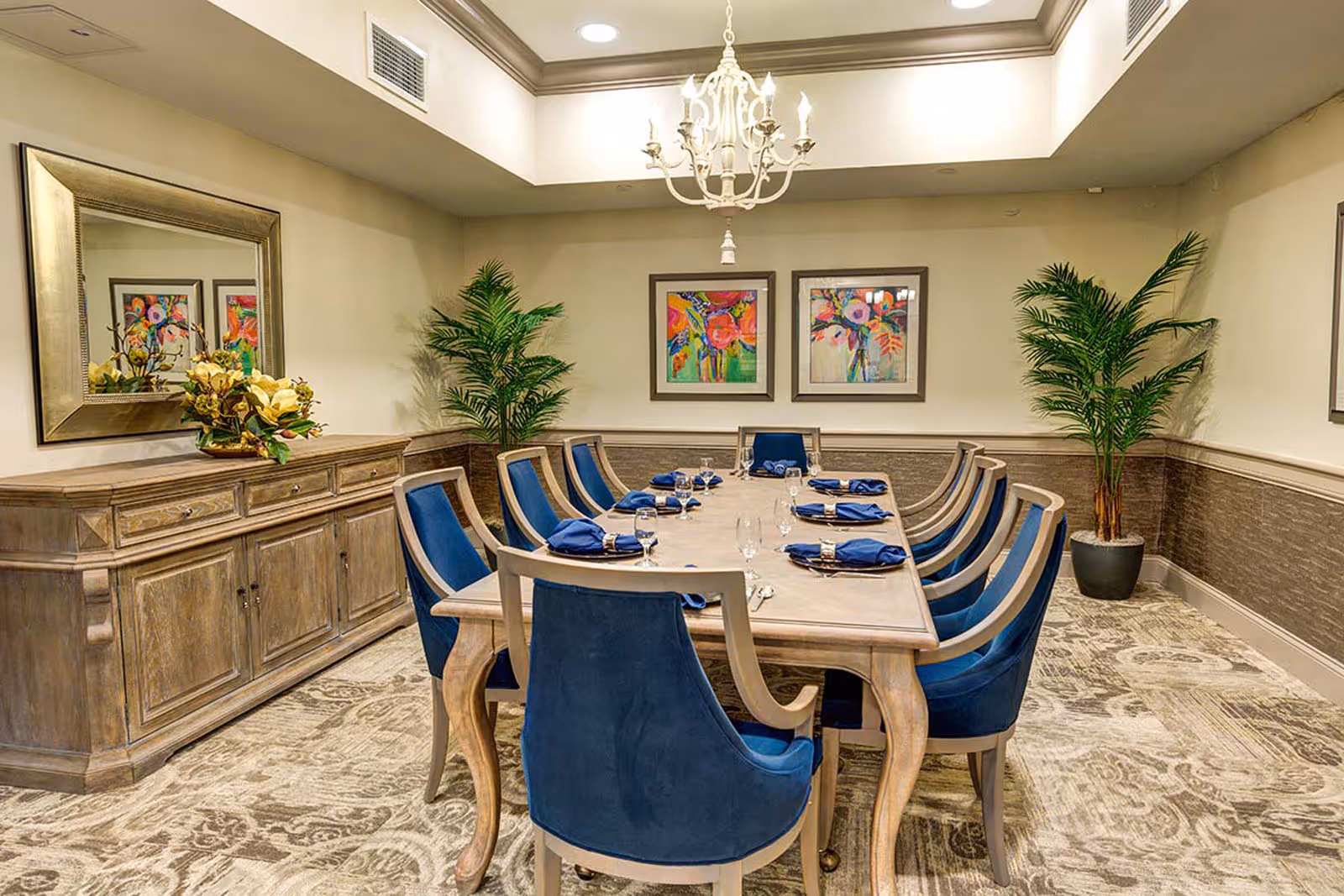 Elegant dining room with a long wooden table set for a meal, blue upholstered chairs, a chandelier, sideboard, and framed artwork on the walls.