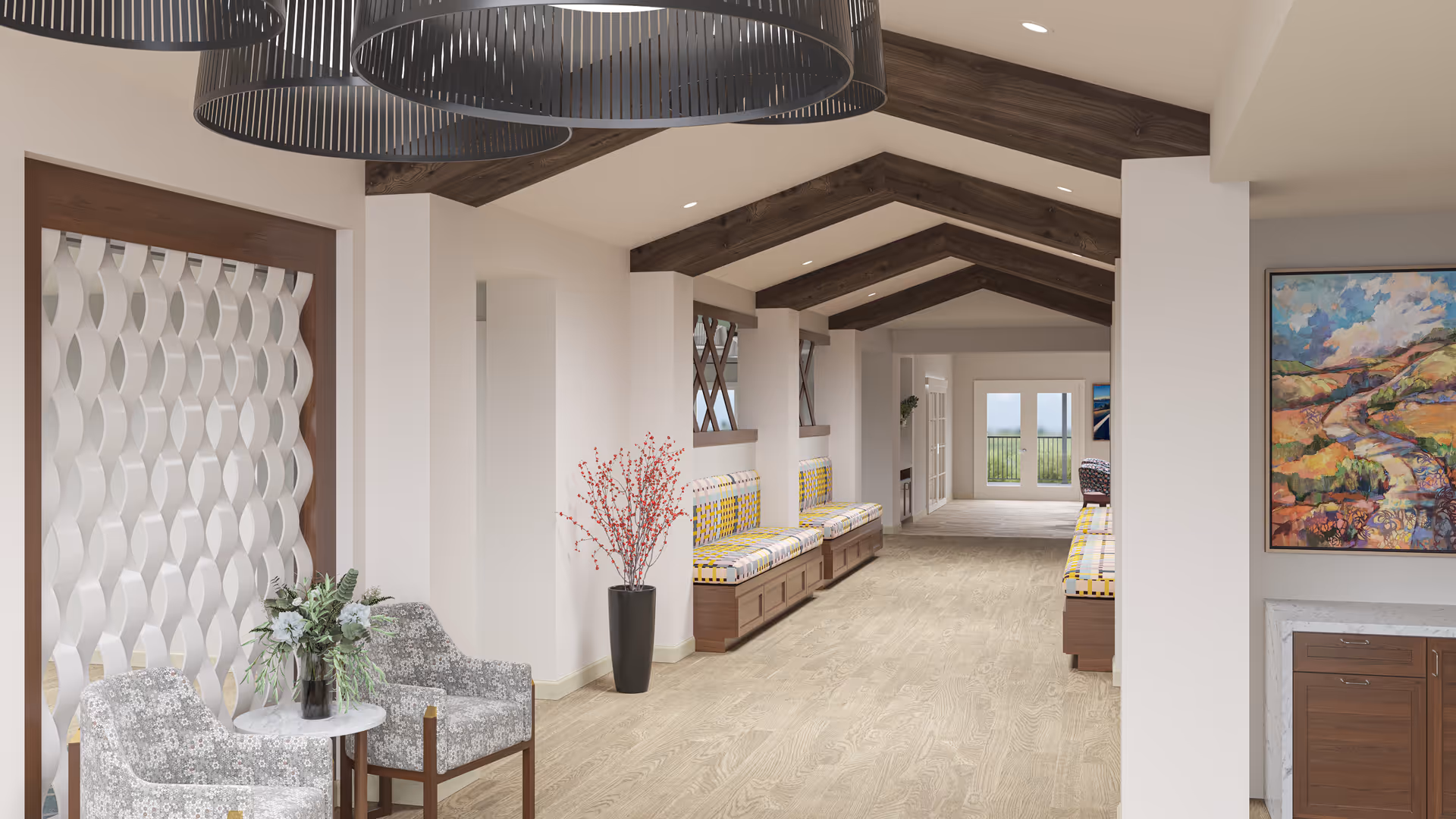 A bright and spacious hallway in Melody Living Independent Living facility featuring wooden ceiling beams, patterned cushioned benches along the walls, two floral upholstered armchairs with a small round table holding a flower arrangement, a tall black vase with red branches, and colorful landscape artwork on the wall. The hallway leads to glass doors with a view outside.