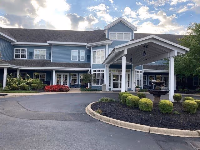 Front exterior view of a two-story senior living facility building with a covered entrance, neatly trimmed bushes, a fountain, and a paved driveway under a partly cloudy sky.