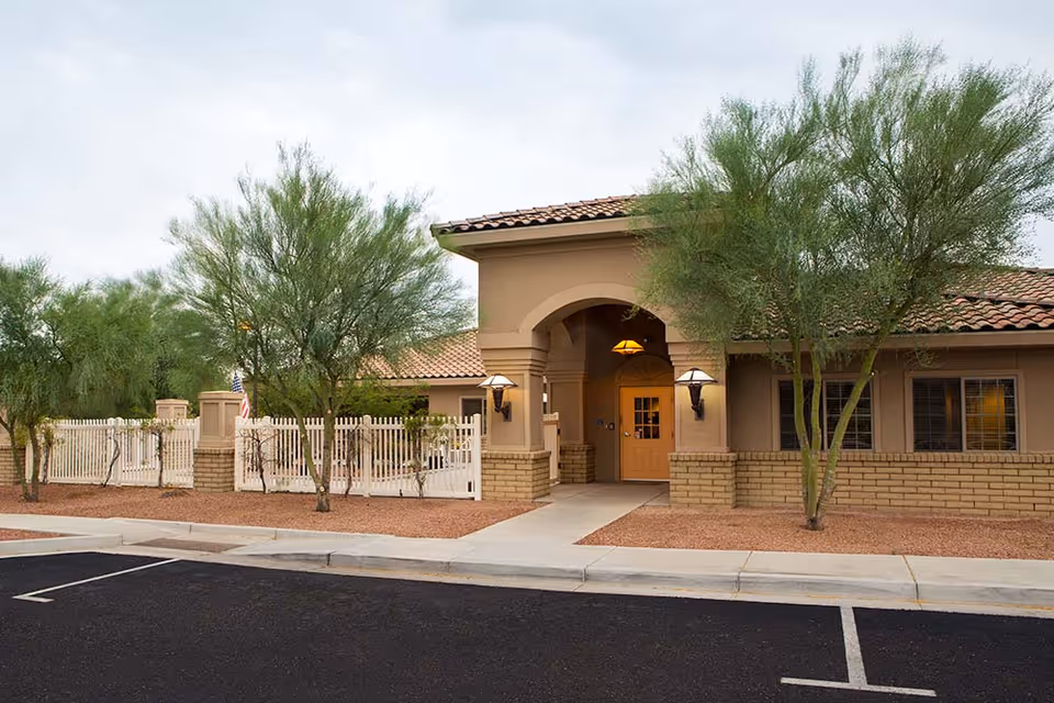 Exterior view of Lifestream At Northeast Phoenix facility showing a single-story building with a tiled roof, beige walls, and a covered entrance with two wall-mounted lamps. There are several green trees and a white fence surrounding the property, with a paved parking area in the foreground.