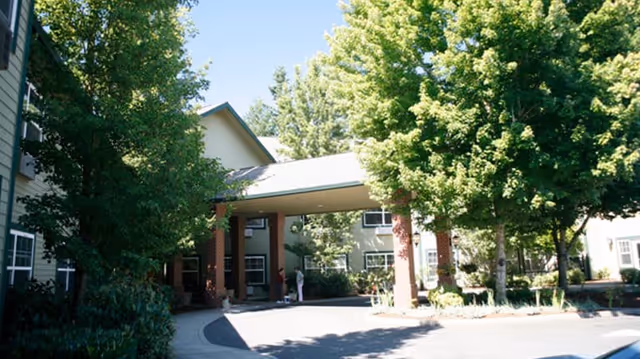 Entrance area of a senior living facility with a covered drop-off zone supported by brick pillars, surrounded by trees and greenery, with the building visible in the background.