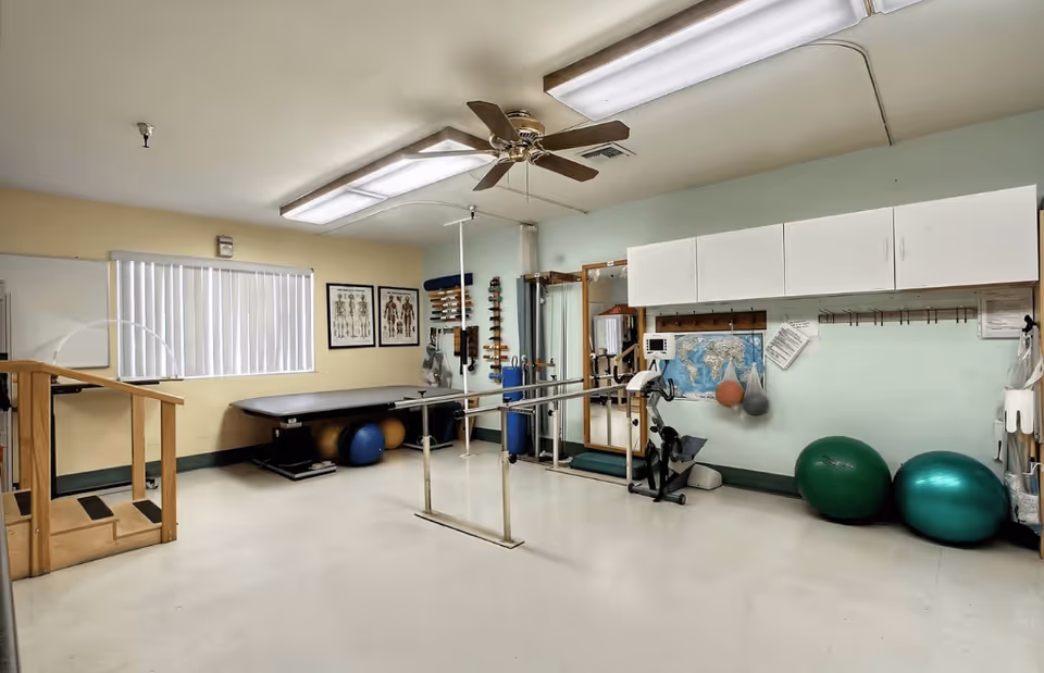 A physical therapy room with parallel bars for walking exercises, exercise balls, a stationary bike, anatomical charts on the wall, and various therapy equipment. The room has a ceiling fan, fluorescent lighting, and a window with vertical blinds.