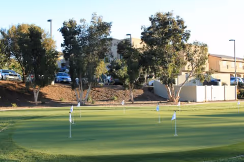 A putting green with several flagsticks in front of trees and a low residential building with parked cars behind it.