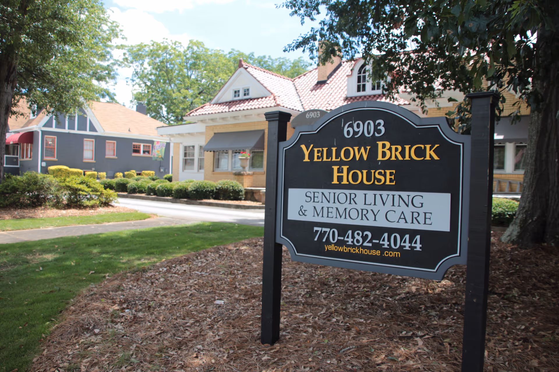 A freestanding sign reading "Yellow Brick House Senior Living & Memory Care" stands in front of a house and landscaped lawn.
