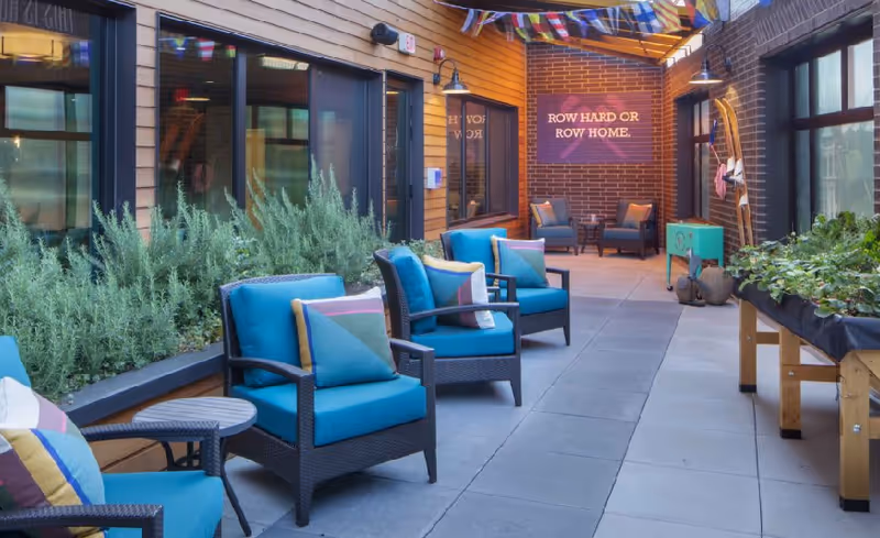 A covered outdoor seating area with blue cushioned chairs and colorful pillows arranged along a walkway. There are plants in raised beds on one side and a table with greenery on the other. At the far end, a brick wall displays a sign that reads 'ROW HARD OR ROW HOME.'