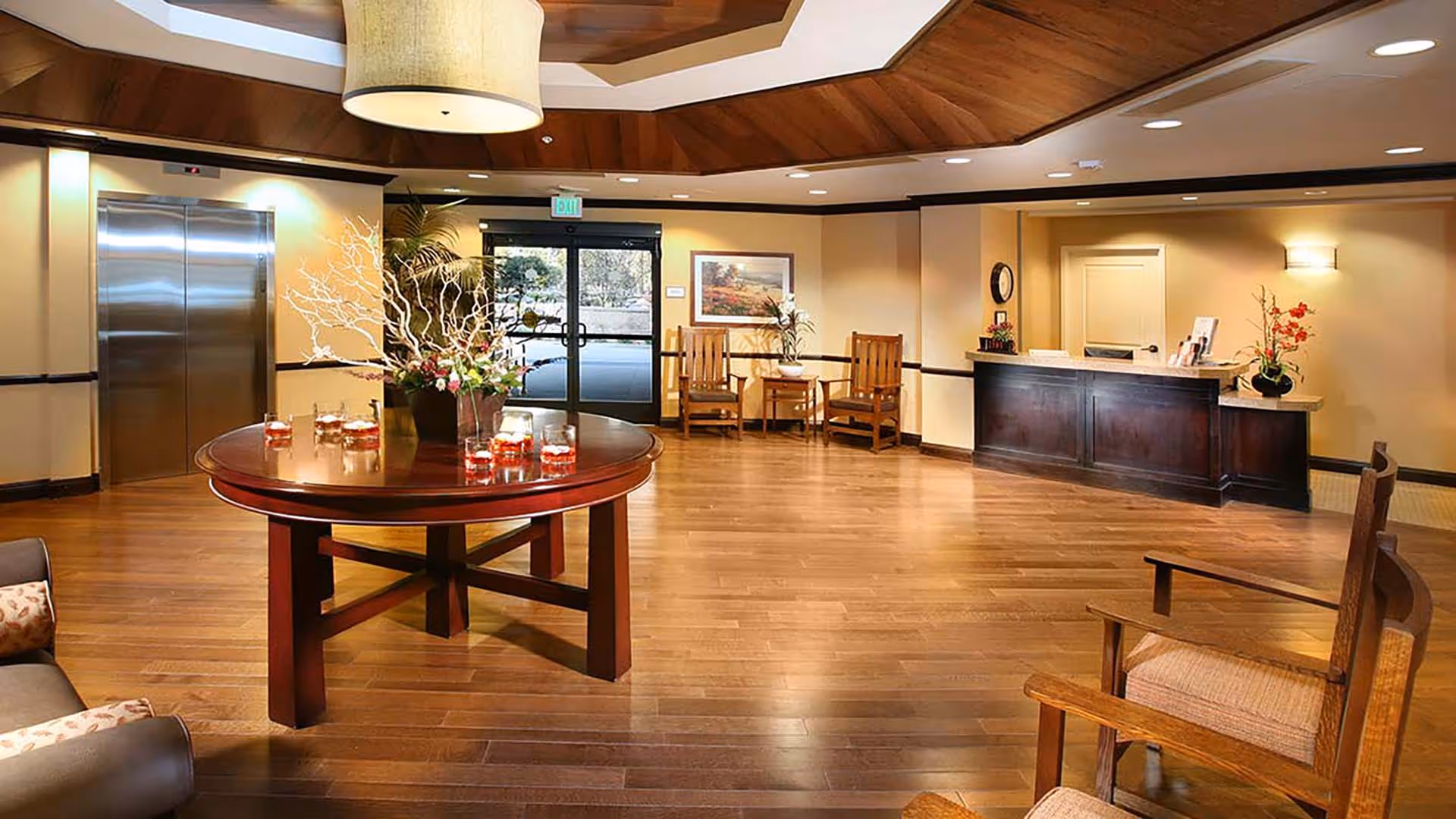 Spacious senior living lobby with a round centerpiece table, seating area, reception desk, elevator and glass entrance doors.