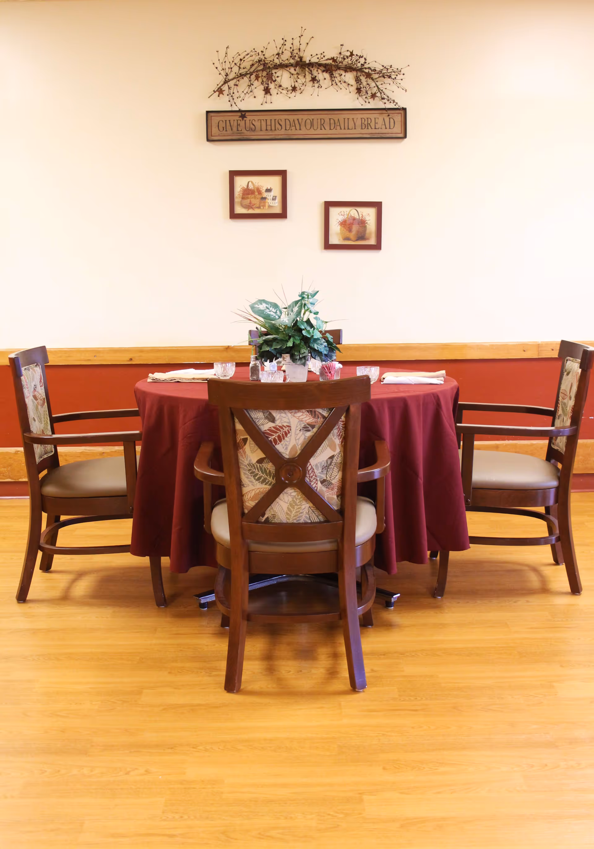A round dining table covered with a burgundy tablecloth, set with four wooden chairs with patterned cushions. The table has a centerpiece of green foliage and is set with glasses and napkins. On the wall behind the table are two framed pictures of baskets and a decorative sign that reads 'GIVE US THIS DAY OUR DAILY BREAD' with a branch decoration above it. The room has wooden flooring and a two-tone wall with a wooden chair rail.