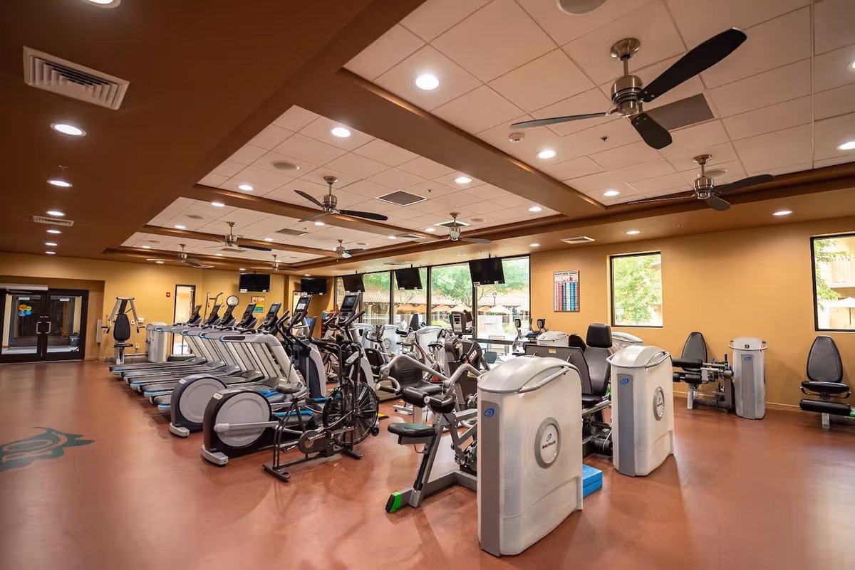 Spacious indoor fitness room with treadmills, stationary bikes, and strength machines under ceiling fans and recessed lighting.