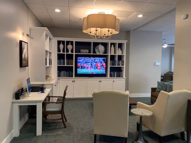 A cozy living room area with a white built-in bookshelf and cabinets, a flat-screen TV displaying a sports game, two beige armchairs, a small round side table, a wooden chair at a white desk with a laptop, and a large ceiling light fixture.