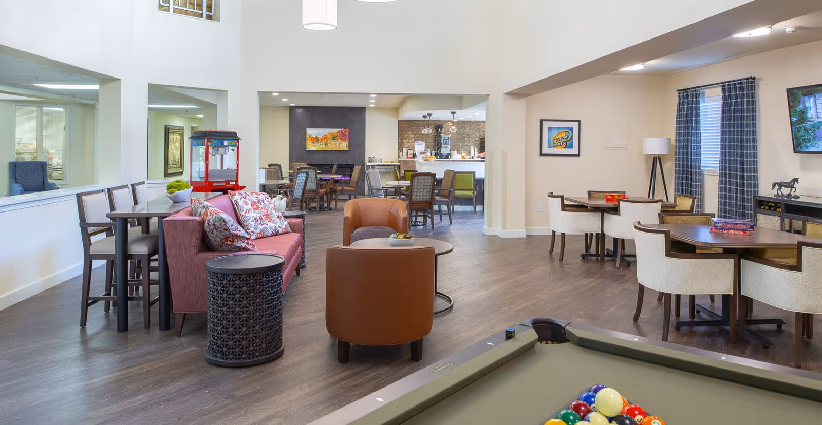 A spacious senior living common area featuring a pool table in the foreground, comfortable seating including a pink sofa with floral pillows and brown chairs, multiple tables with chairs for games or dining, a popcorn machine, and a TV mounted on the wall. The room has wood flooring, large windows with blue checkered curtains, and bright lighting.