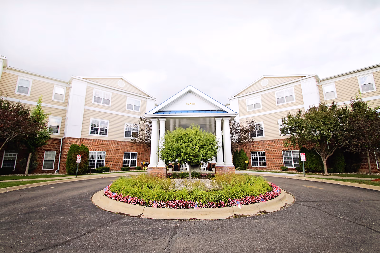 Front exterior view of a senior living facility named Lockwood of Clinton, featuring a circular driveway with a landscaped roundabout in the center, a covered entrance with white columns, and a three-story building with beige siding and red brick on the lower level.