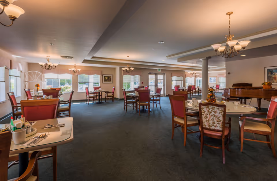 Spacious dining room with multiple round and rectangular tables, red upholstered chairs, chandeliers, and a piano.