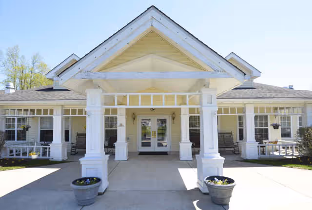 Covered entrance of a single-story senior living facility featuring white columns, rocking chairs, and potted planters.