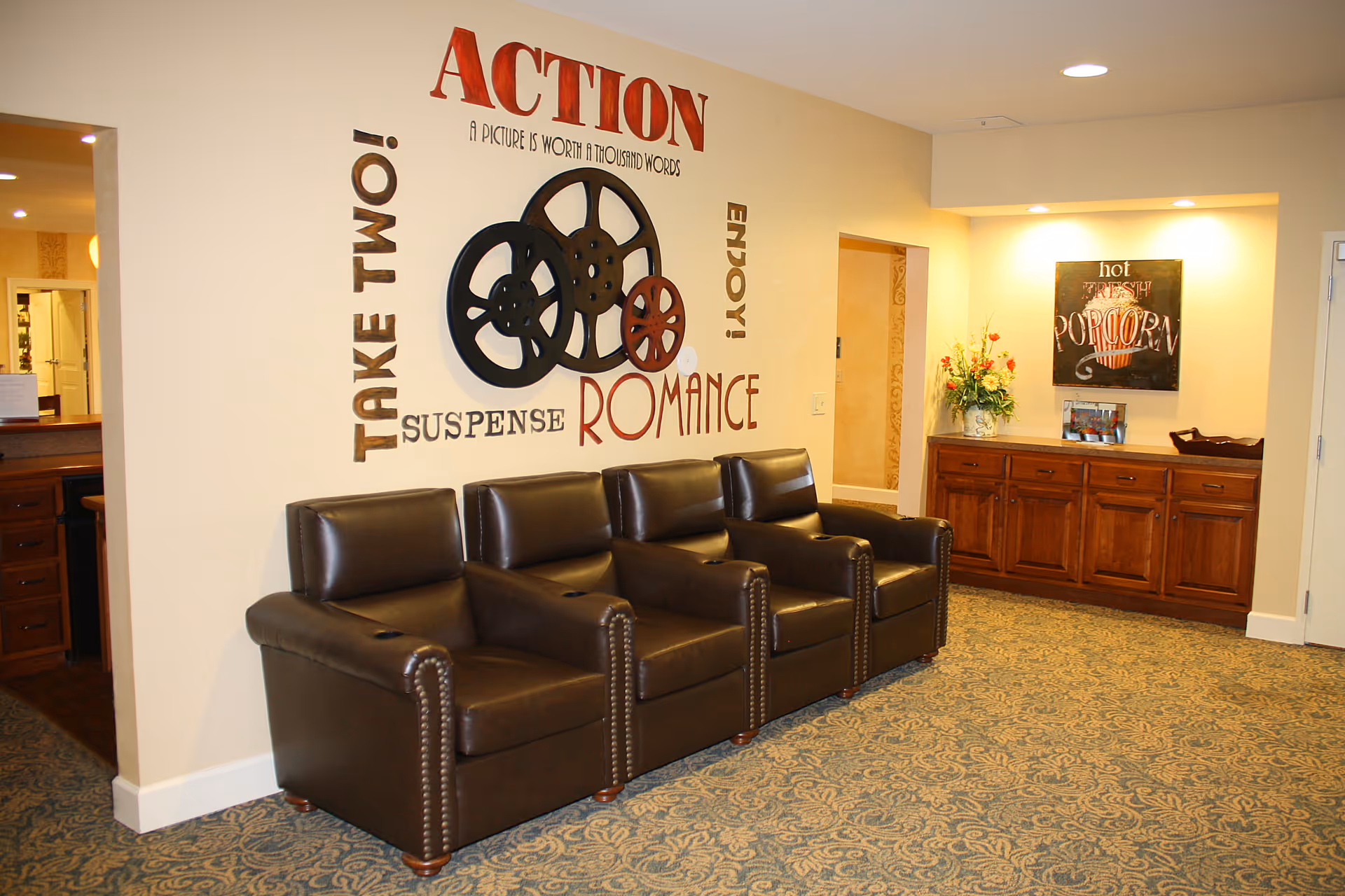 A cozy seating area with four brown leather armchairs arranged in a row against a wall. The wall features decorative film reels and words related to movies such as 'ACTION', 'TAKE TWO!', 'SUSPENSE', 'ROMANCE', and 'ENJOY!'. To the right, there is a wooden cabinet with a flower arrangement and a sign that reads 'hot FRESH POPCORN'. The carpet has a blue and beige pattern, and the lighting is warm and inviting.