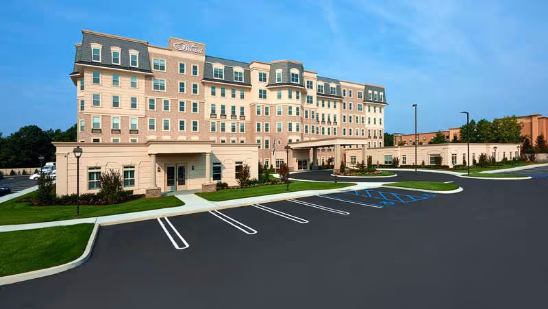 Front exterior of The Bristal at Garden City senior living building with a large empty parking lot under a clear blue sky.