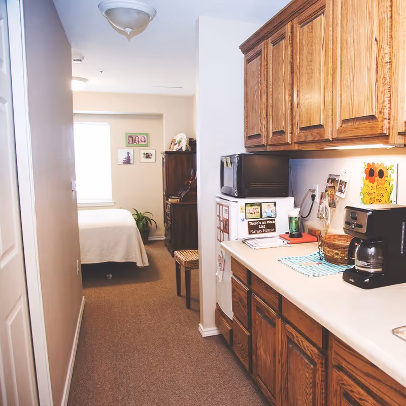 A narrow interior space showing a small kitchenette area with wooden cabinets, a coffee maker, a microwave on top of a mini refrigerator, and various personal items and photos on the counter and walls. Beyond the kitchenette is a bedroom with a bed covered in a white blanket, a plant near the window, and framed pictures on the wall.