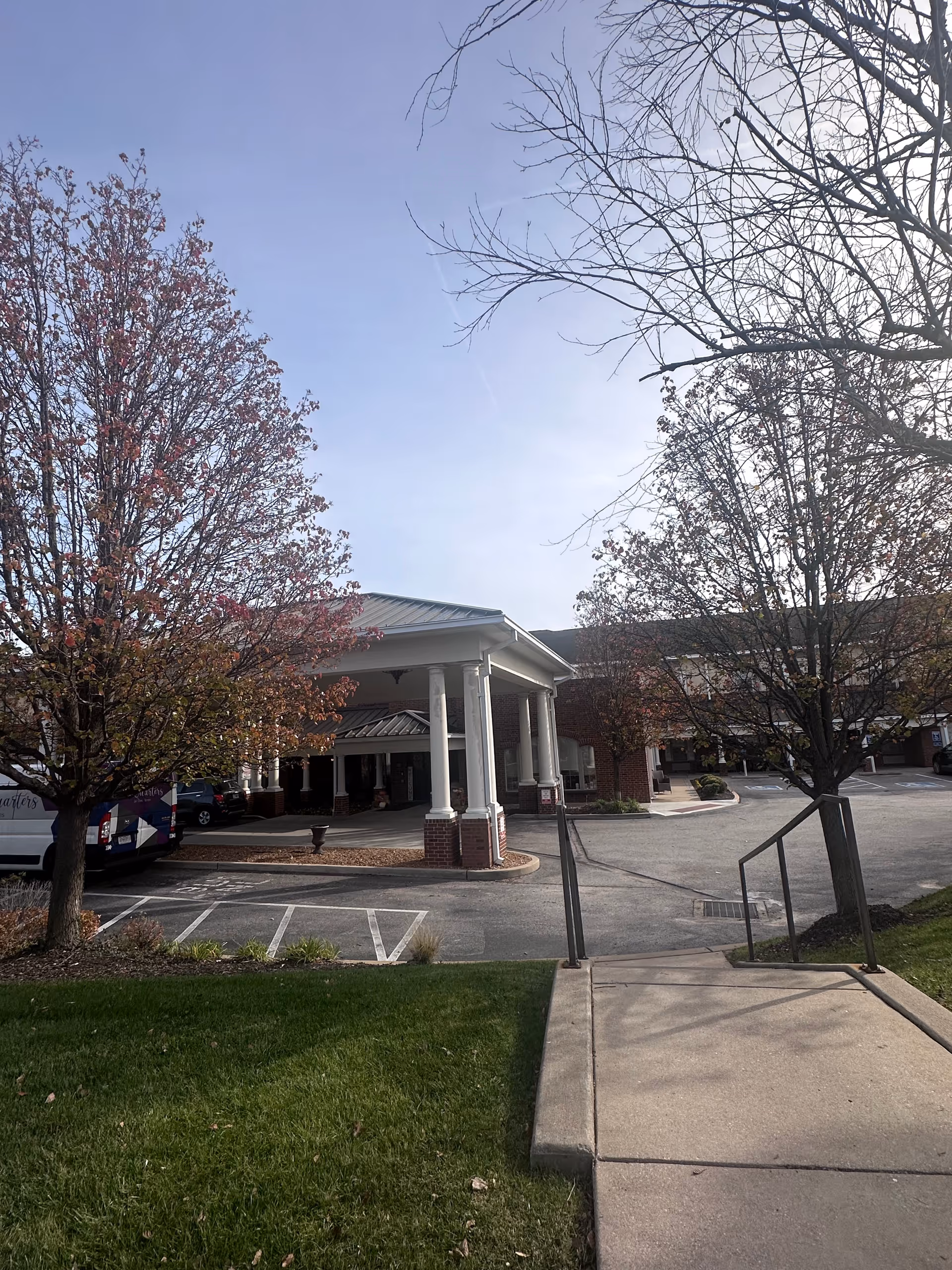 Exterior view of the entrance to a senior living facility named Quarters At Des Peres, showing a covered drop-off area with white columns and a brick base. There are trees with autumn foliage on either side of the driveway, a concrete walkway with handrails leading up to the entrance, and a parked van partially visible on the left.