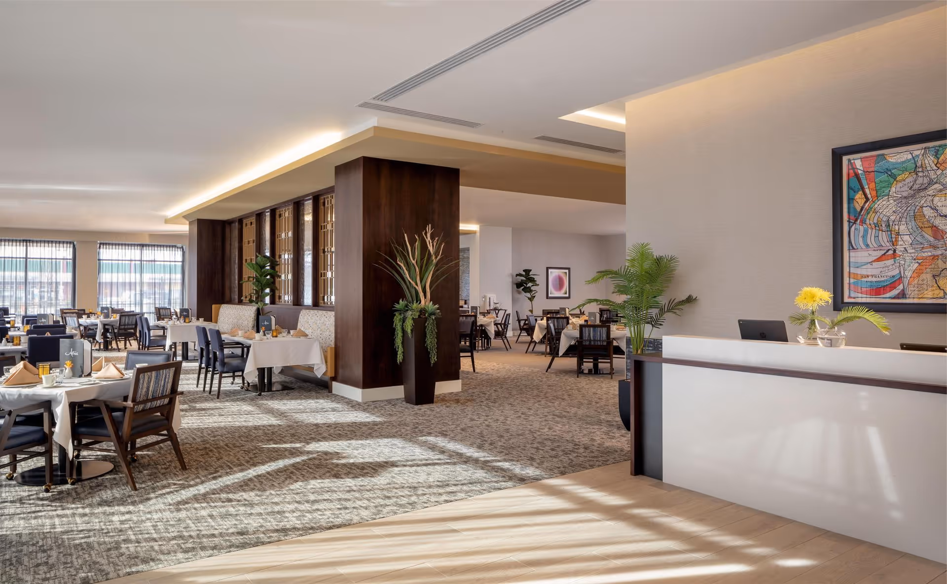 A spacious and well-lit dining room with multiple tables set with white tablecloths, napkins, and tableware. The room features large windows with sheer curtains allowing natural light to fill the space. There are dark wooden chairs and decorative plants placed around the room. A reception desk with a flower vase and a colorful abstract painting on the wall is visible in the foreground.