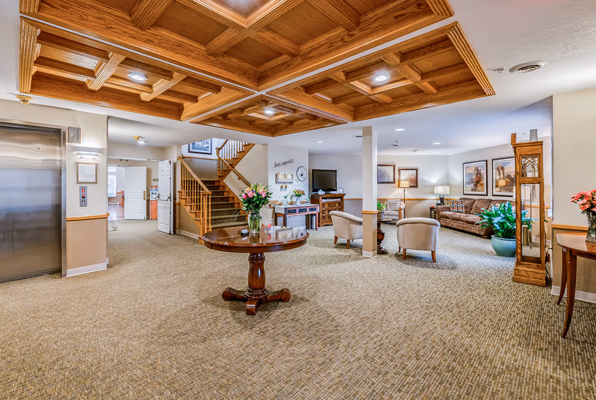 A spacious and well-lit common area in a senior living facility featuring a wooden coffered ceiling with recessed lighting, a round wooden table with a vase of flowers in the center, an elevator on the left, carpeted floors, a staircase with wooden railings, and a seating area with armchairs, a sofa, framed artwork, plants, and a television.