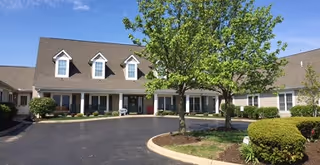 Exterior view of a senior living community building with a paved driveway, landscaped greenery including a tree and bushes, and a clear blue sky.