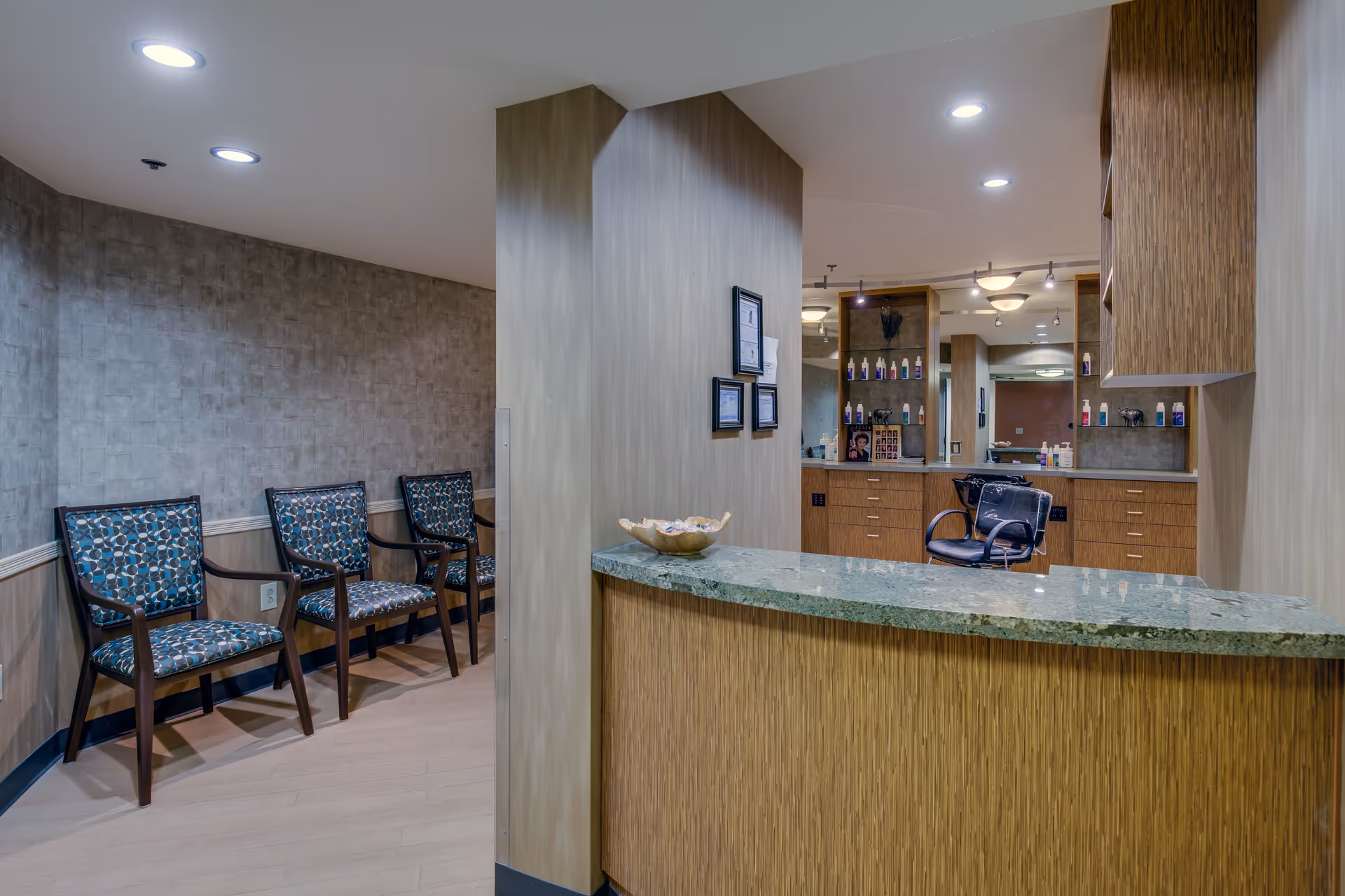 Interior view of a salon area in a senior living facility with a green marble countertop reception desk, wooden cabinetry, a black salon chair, and shelves with hair care products. To the left, there are three patterned chairs lined up against a textured wall under recessed ceiling lights.