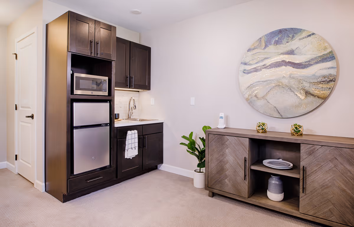 A small kitchenette area with dark wood cabinets, a microwave, a mini refrigerator, and a sink with a towel hanging on the cabinet handle. Next to the kitchenette is a wooden sideboard with decorative items including two small plants, a plate, and a vase. Above the sideboard is a large round abstract wall art. The floor is carpeted and the walls are painted light beige.