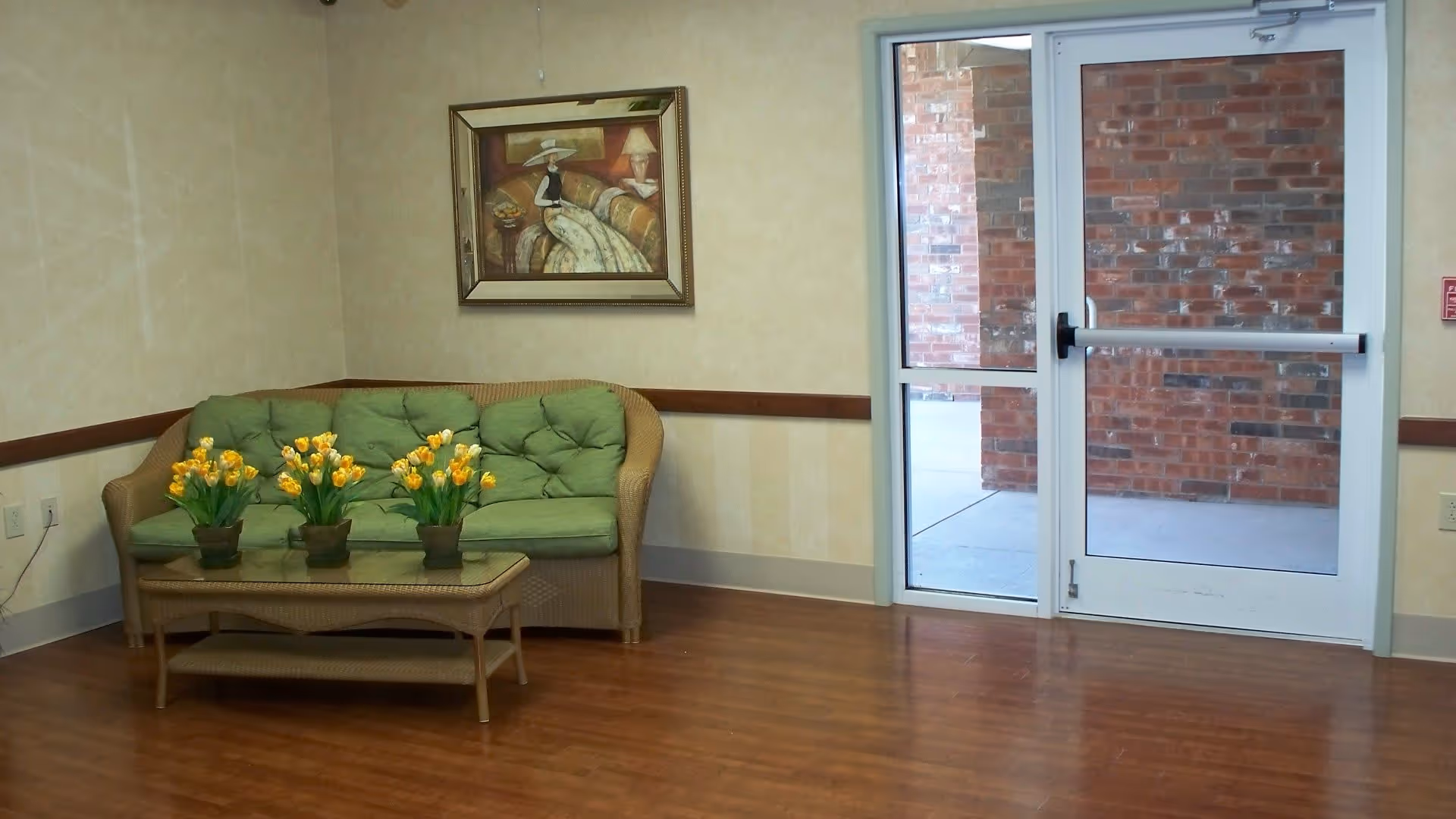 A small seating area with a wicker couch featuring green cushions and a glass-top wicker coffee table holding three pots of yellow tulips. A framed painting of a woman in a hat sitting on a couch hangs on the wall above the couch. To the right, there is a glass door leading outside, showing a brick wall and concrete ground.