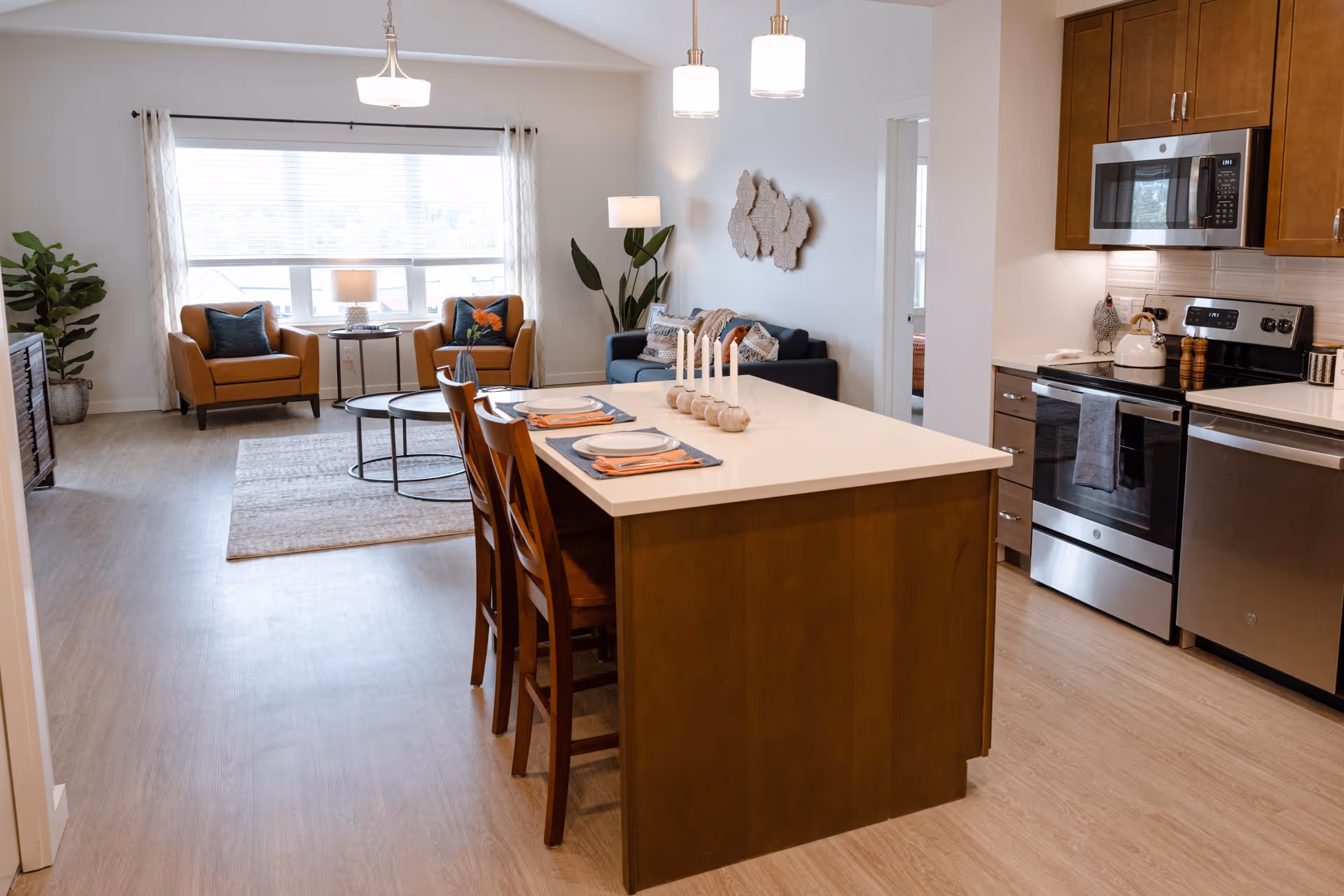 Open concept living space with a kitchen island featuring four place settings and decorative candles. The kitchen has wooden cabinets, a stainless steel stove, microwave, and dishwasher. The living area includes a dark blue sofa, two orange armchairs, a coffee table, a floor lamp, and a large window with white curtains letting in natural light.
