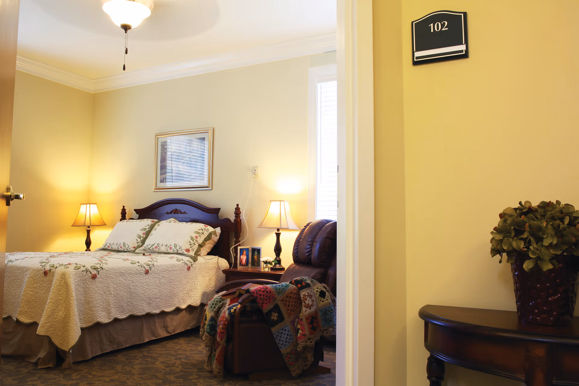 Well-lit bedroom with a made bed, two bedside lamps, an armchair draped with a crocheted blanket, a small table with a plant, and a '102' room sign on the wall.
