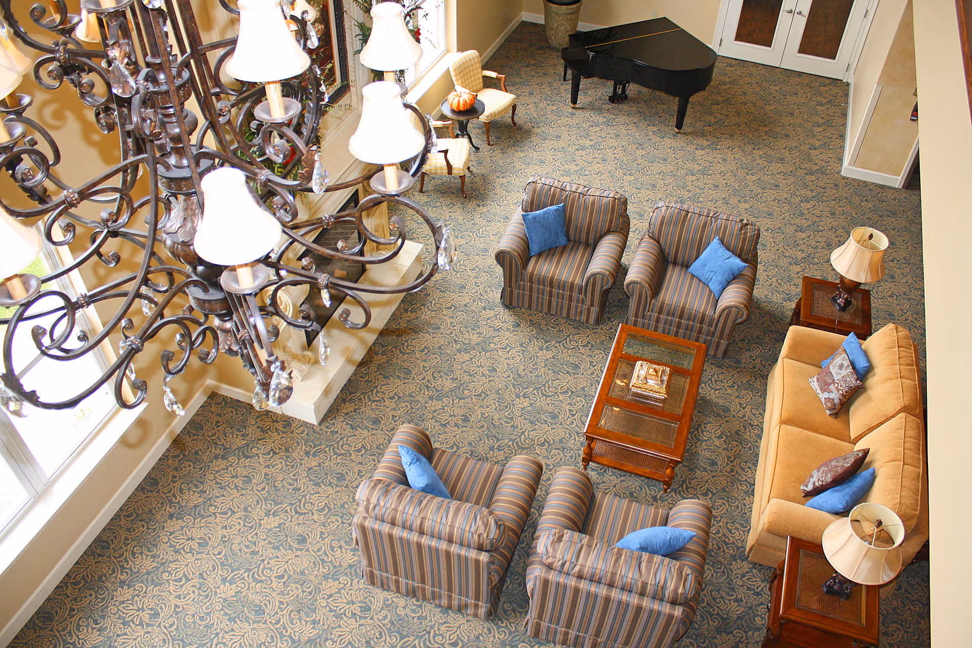 Overhead view of a living room area with a patterned carpet, a large chandelier, a fireplace, a grand piano, a beige sofa with pillows, four striped armchairs with blue pillows, two side tables with lamps, and two small chairs with a small round table holding a pumpkin.
