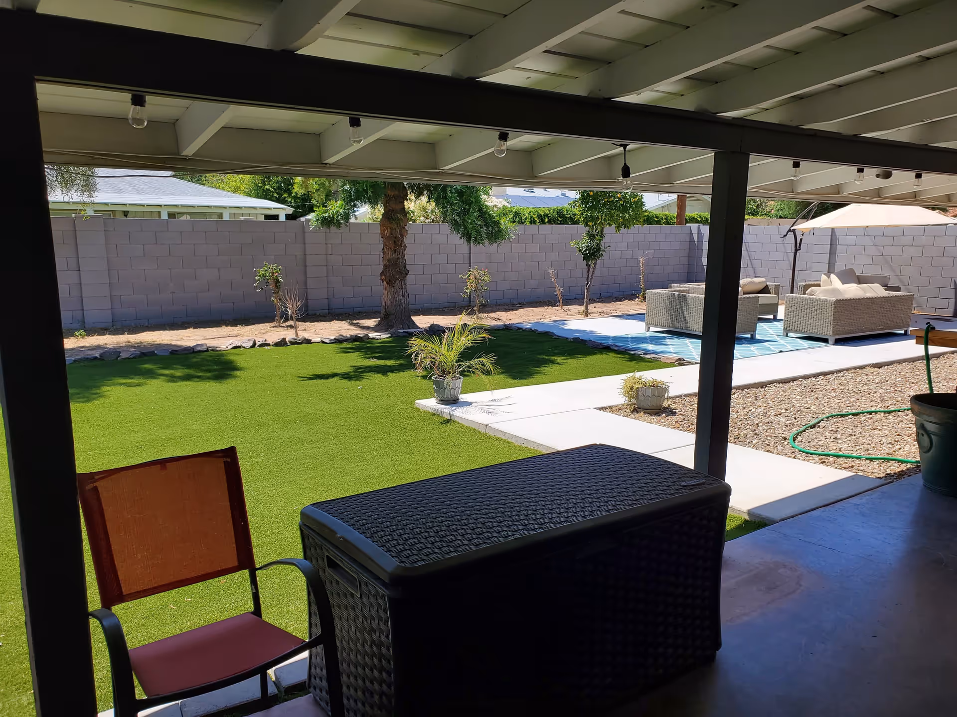 View from a covered patio looking out onto a backyard with artificial grass, small plants, and a seating area with outdoor furniture and an umbrella. The yard is enclosed by a gray brick wall.