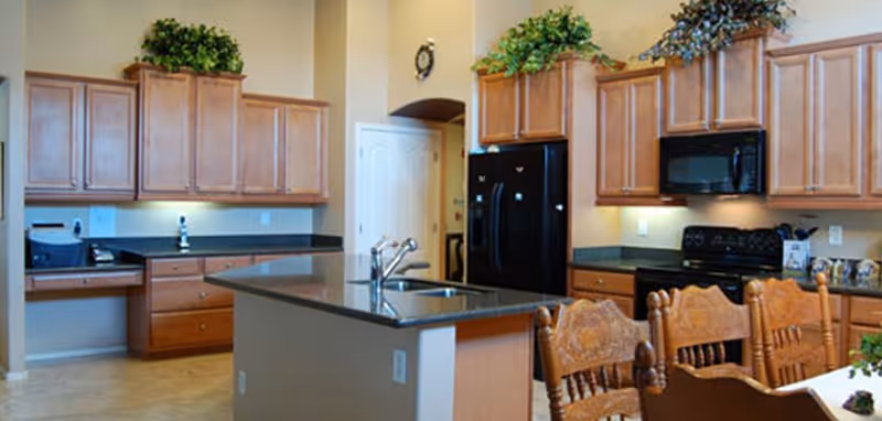 A spacious kitchen with wooden cabinets, a black refrigerator, black microwave, black stove, and a central island with a sink. There are decorative plants on top of the cabinets and wooden chairs around a dining table in the foreground.
